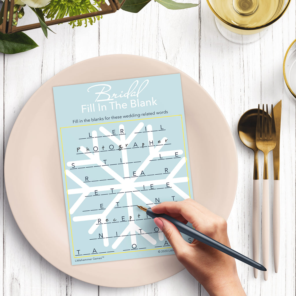 Woman’s hand holding a pen playing a Scandinavian snowflake design Bridal Fill in the Blank game card at a light place setting