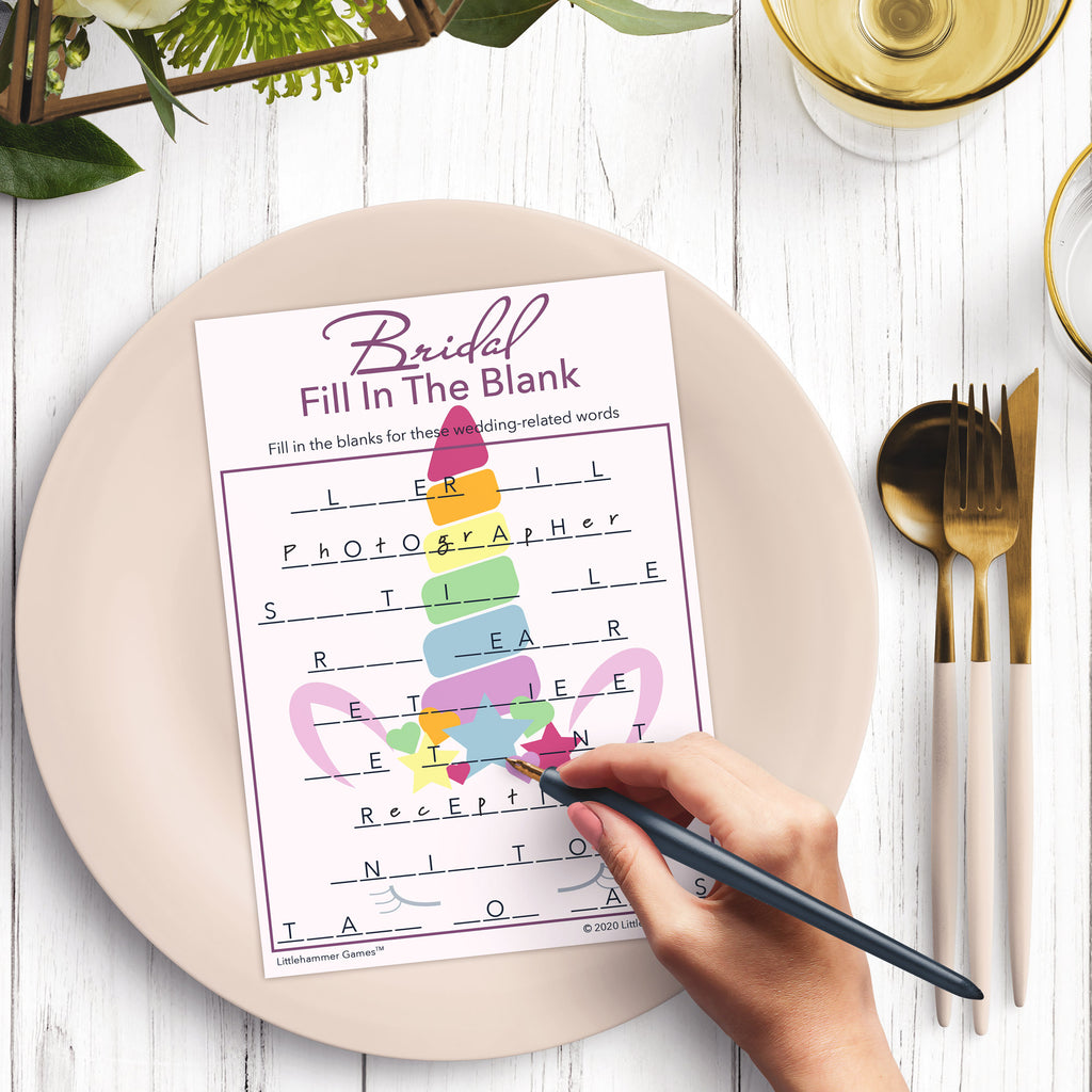 Woman’s hand holding a pen playing a pastel rainbow unicorn Bridal Fill in the Blank game card on a pink background at a light place setting