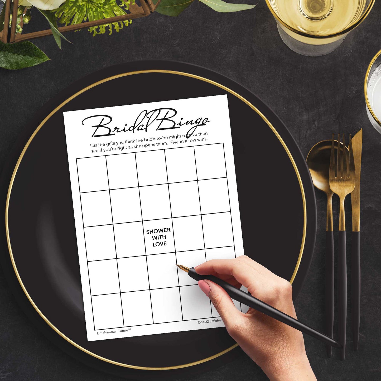 Woman with a pen sitting at a dark place setting with a black and gold plate filling out a minimalist black and white Bridal Gift Bingo card