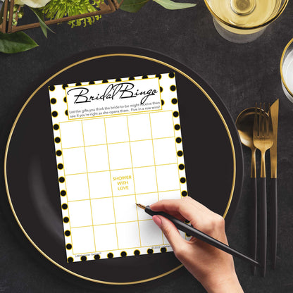 Woman with a pen sitting at a dark place setting with a black and gold plate filling out a black and gold polka dot Bridal Gift Bingo card