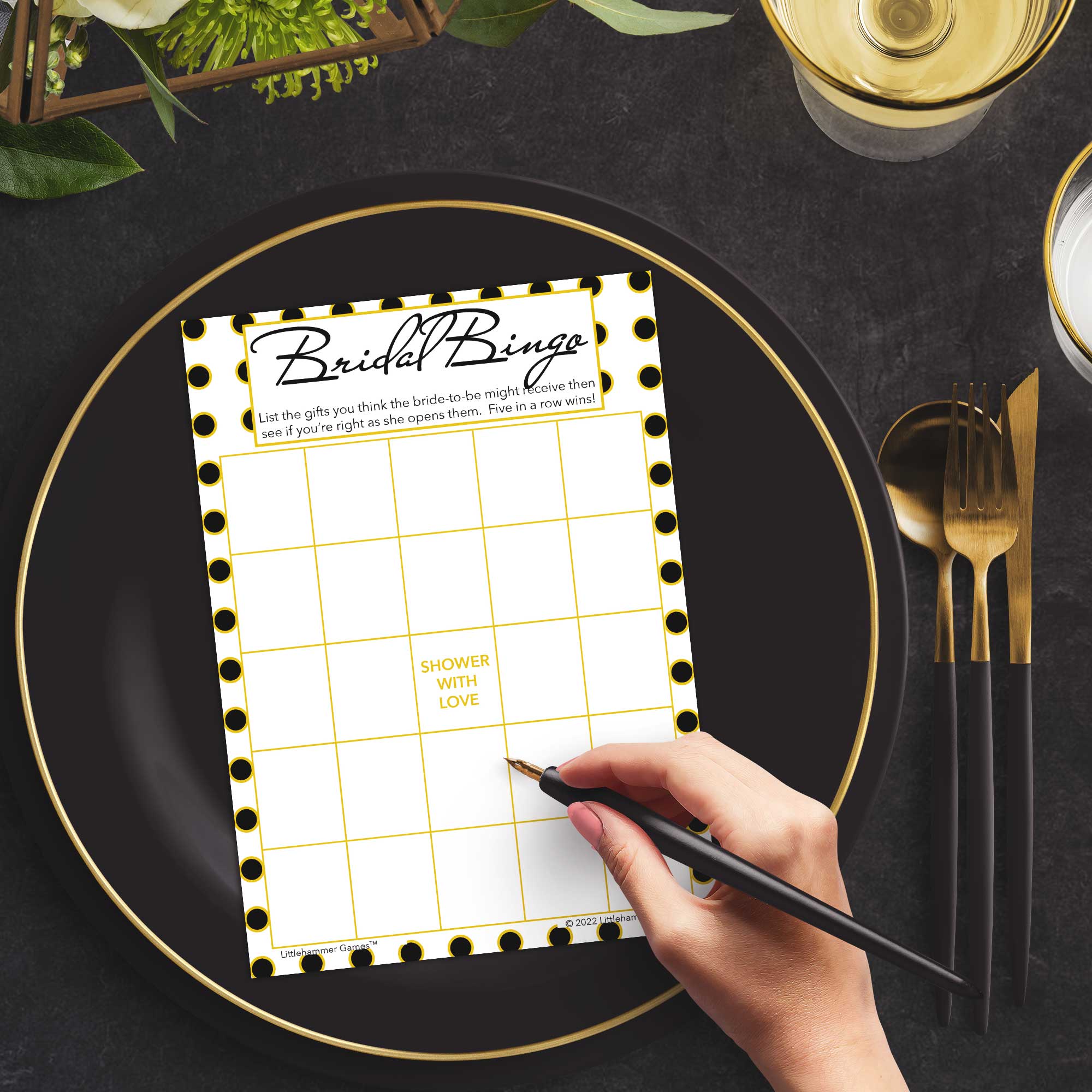 Woman with a pen sitting at a dark place setting with a black and gold plate filling out a black and gold polka dot Bridal Gift Bingo card