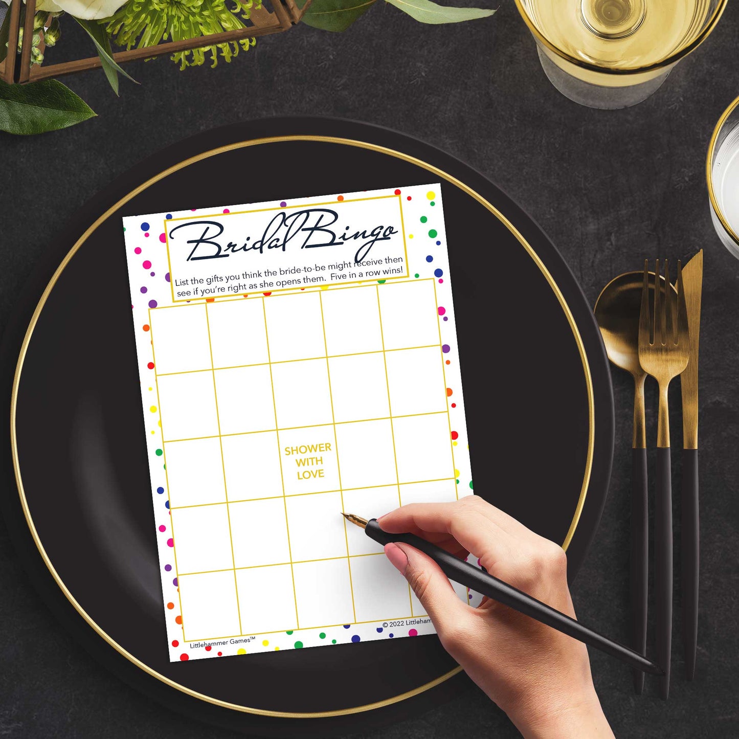 Woman with a pen sitting at a dark place setting with a black and gold plate filling out a rainbow polka dot Bridal Gift Bingo card