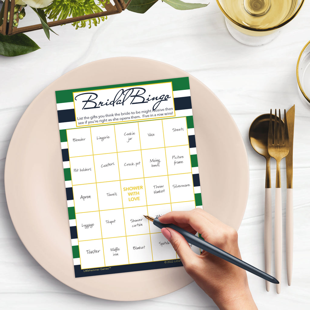 Woman’s hand holding a pen playing a green and navy stripe Bridal Gift Bingo game card at a light place setting