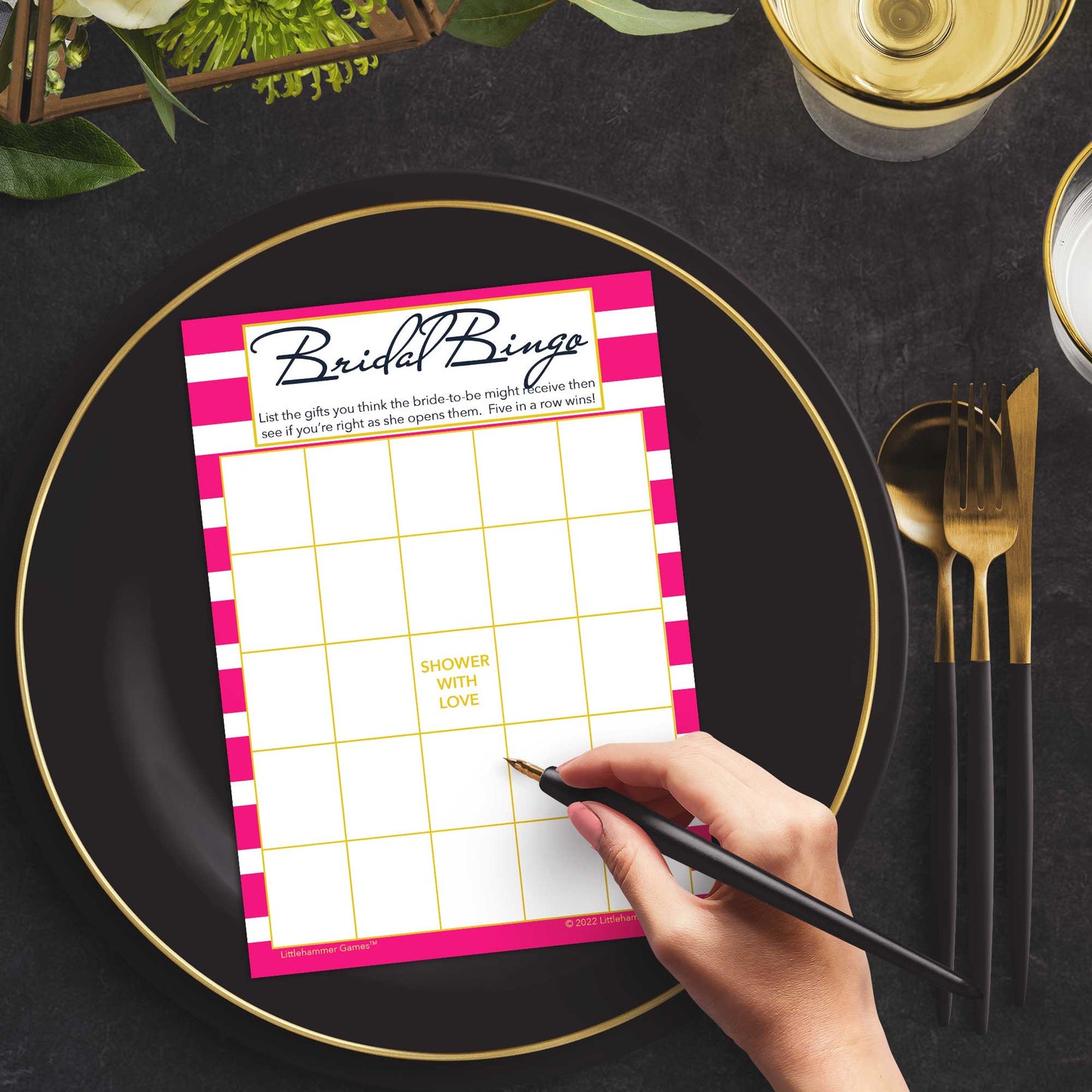 Woman with a pen sitting at a dark place setting with a black and gold plate filling out a hot pink-striped Bridal Gift Bingo card