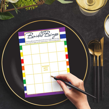 Woman with a pen sitting at a dark place setting with a black and gold plate filling out a rainbow-striped Bridal Gift Bingo card