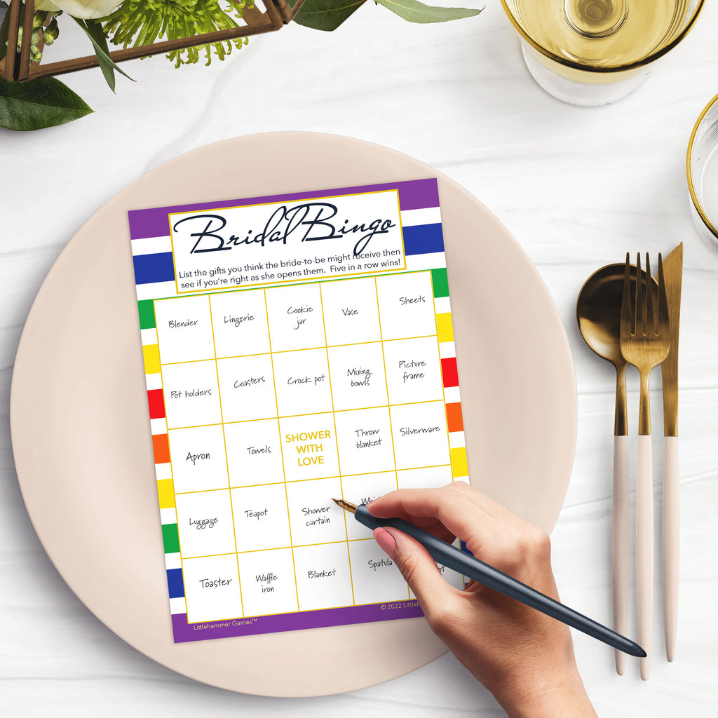 Woman with a pen sitting at a dark place setting with a black and gold plate filling out a rainbow-striped Bridal Gift Bingo card