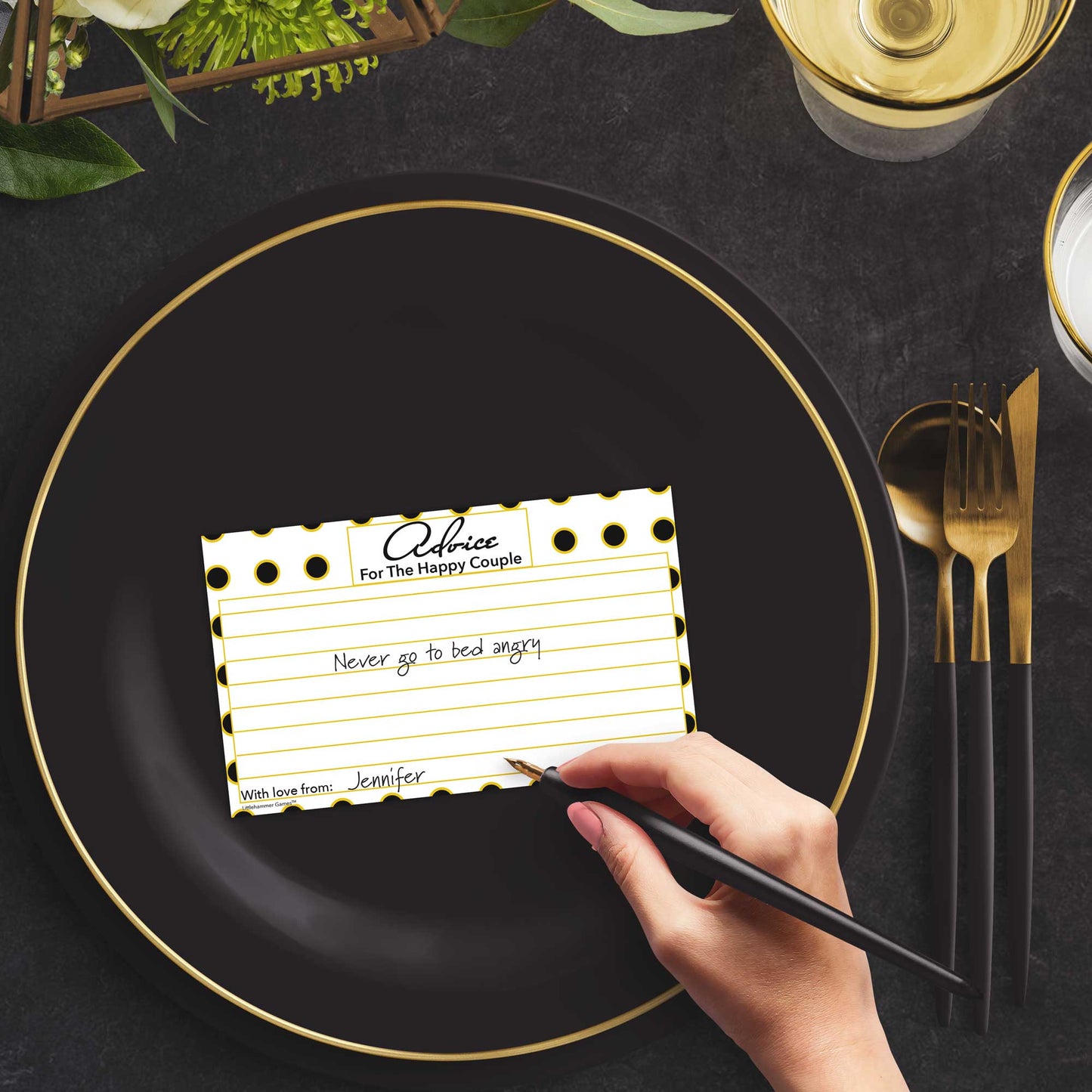 Woman with a pen sitting at a dark place setting with a black and gold plate filling out a black and gold polka dot Advice for the Happy Couple card