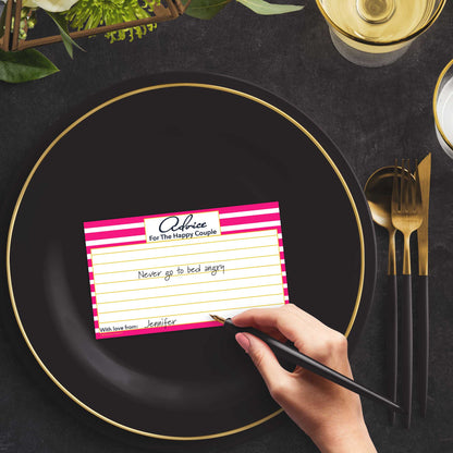 Woman with a pen sitting at a dark place setting with a black and gold plate filling out a pink-striped Advice for the Happy Couple card