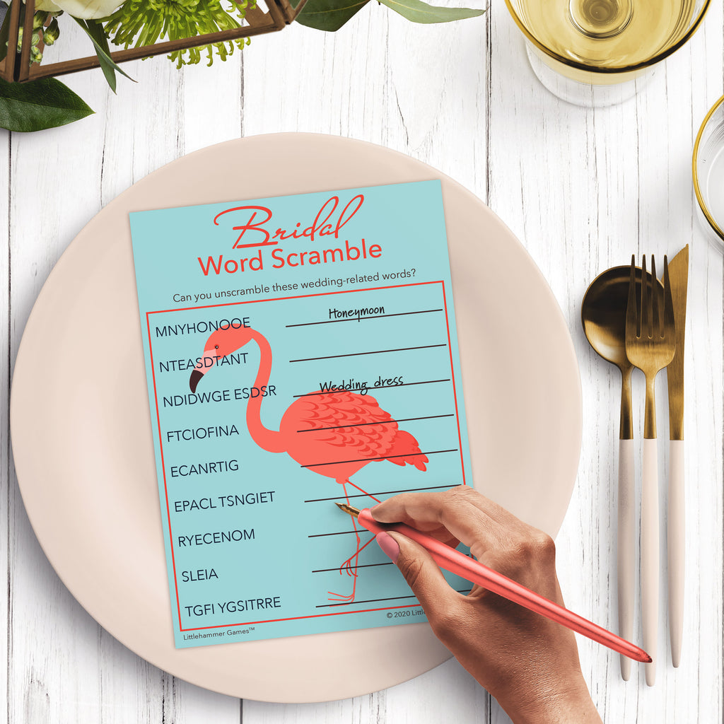 Woman’s hand holding a pen playing a flamingo-themed Bridal Word Scramble game card at a light place setting