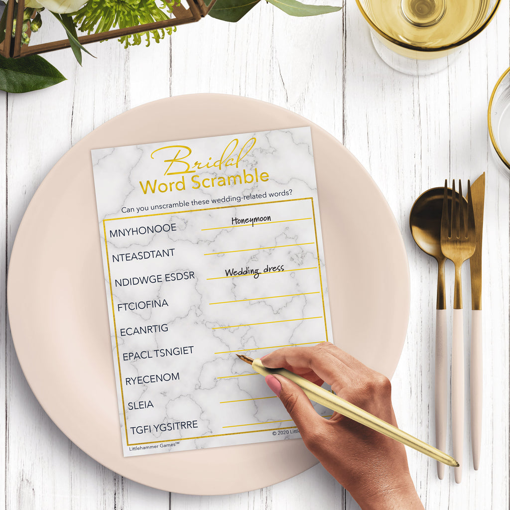 Woman’s hand holding a pen playing a gold marbled Bridal Word Scramble game card at a light place setting