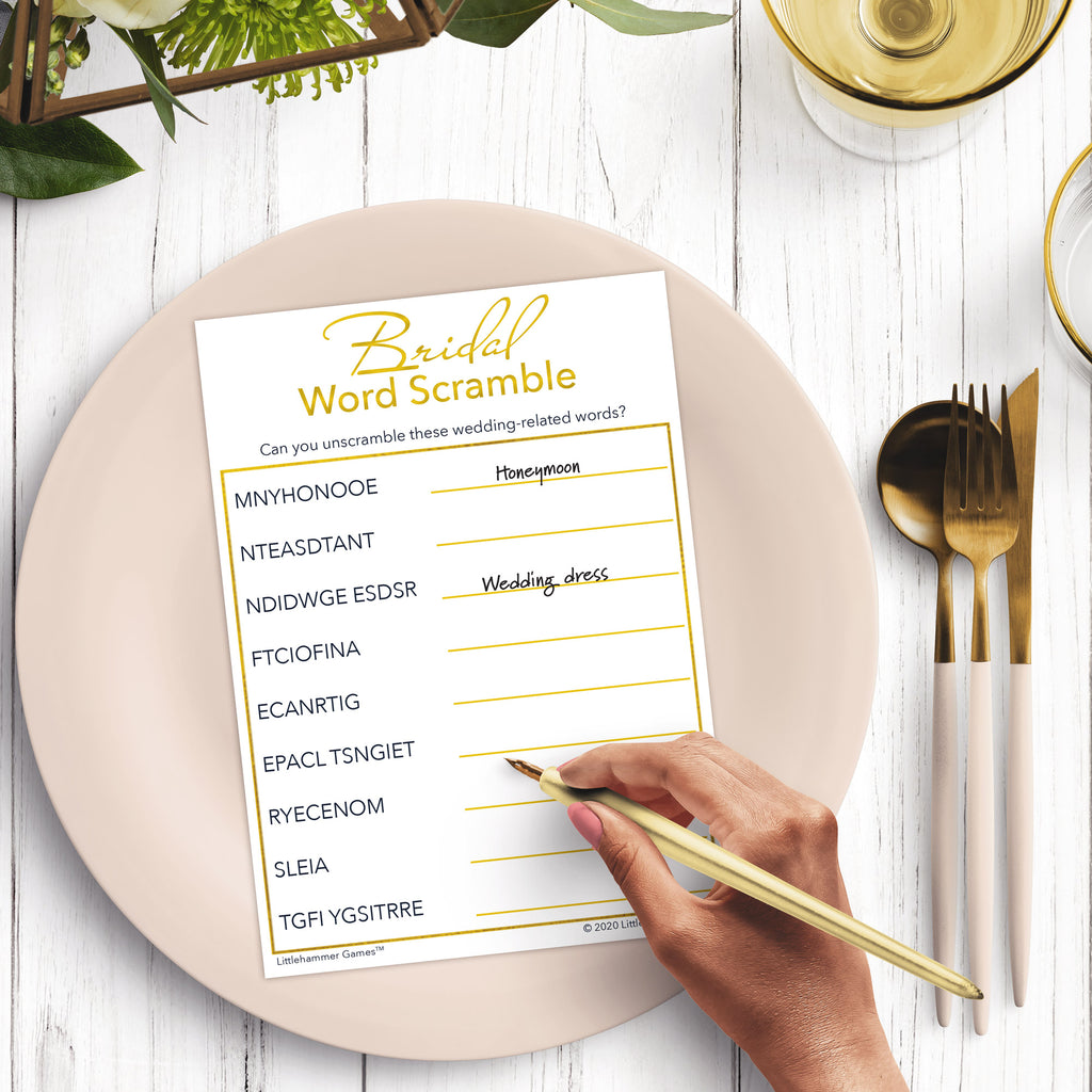 Woman’s hand holding a pen playing a gold and white Bridal Word Scramble game card at a light place setting