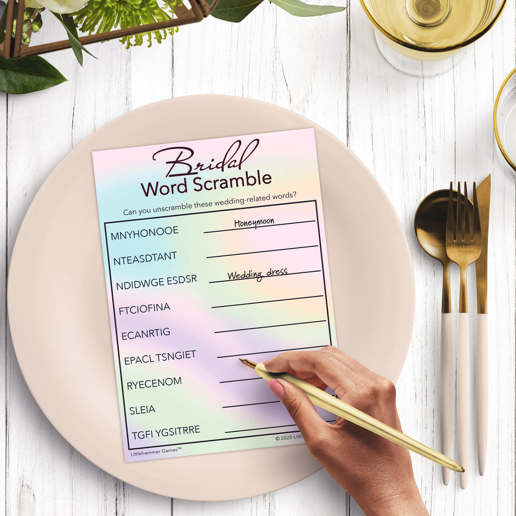 Woman’s hand holding a pen playing a holographic-themed Bridal Word Scramble game card at a light place setting