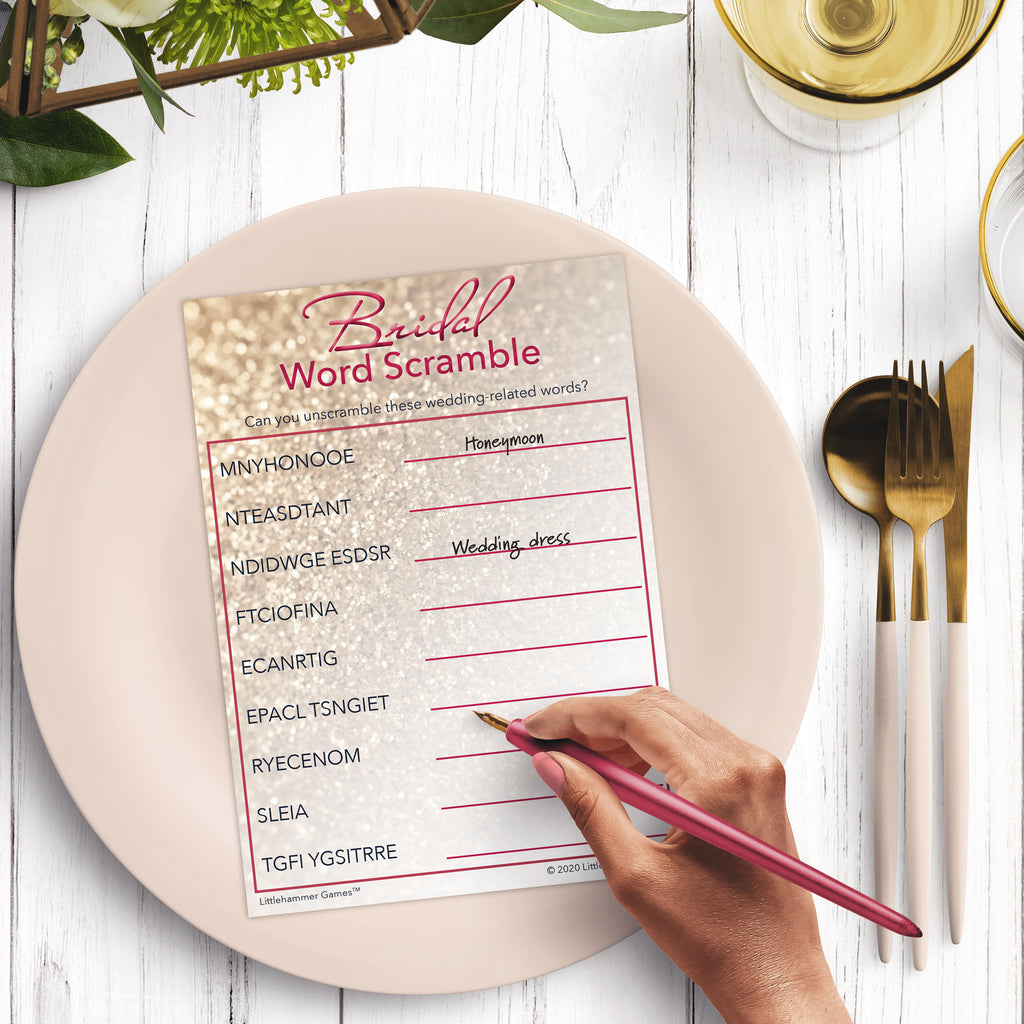 Woman’s hand holding a pen playing a glittery rose gold Bridal Word Scramble game card at a light place setting
