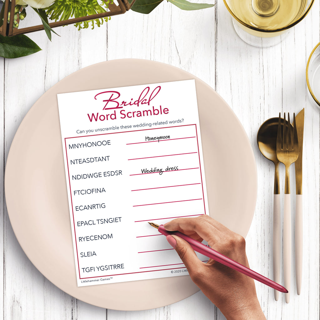 Woman’s hand holding a pen playing a white and rose gold Bridal Word Scramble game card at a light place setting