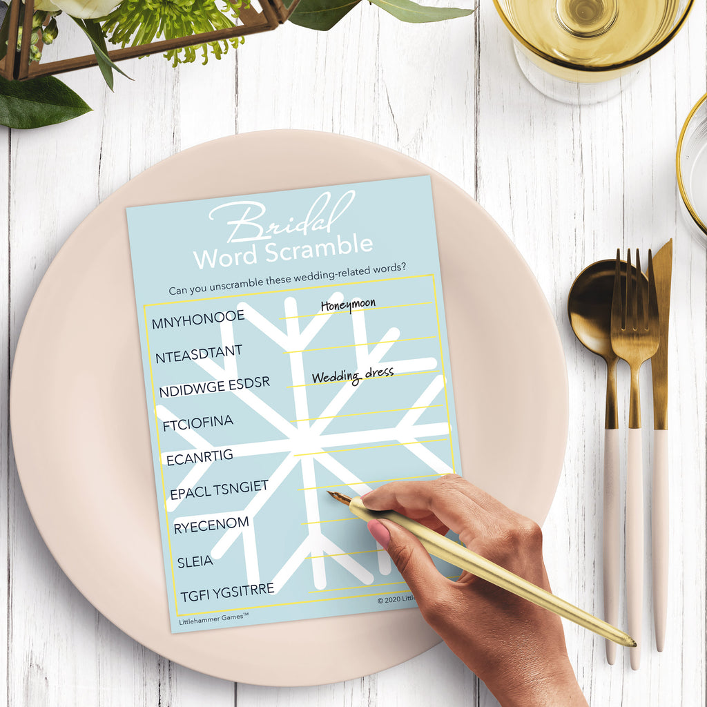 Woman’s hand holding a pen playing a snowflake Bridal Word Scramble game card at a light place setting