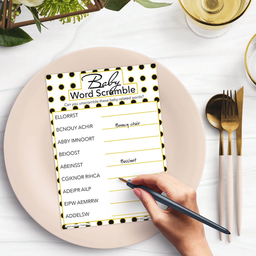 Woman’s hand holding a pen playing a geometric black and gold polka dot Baby Word Scramble game card at a light place setting