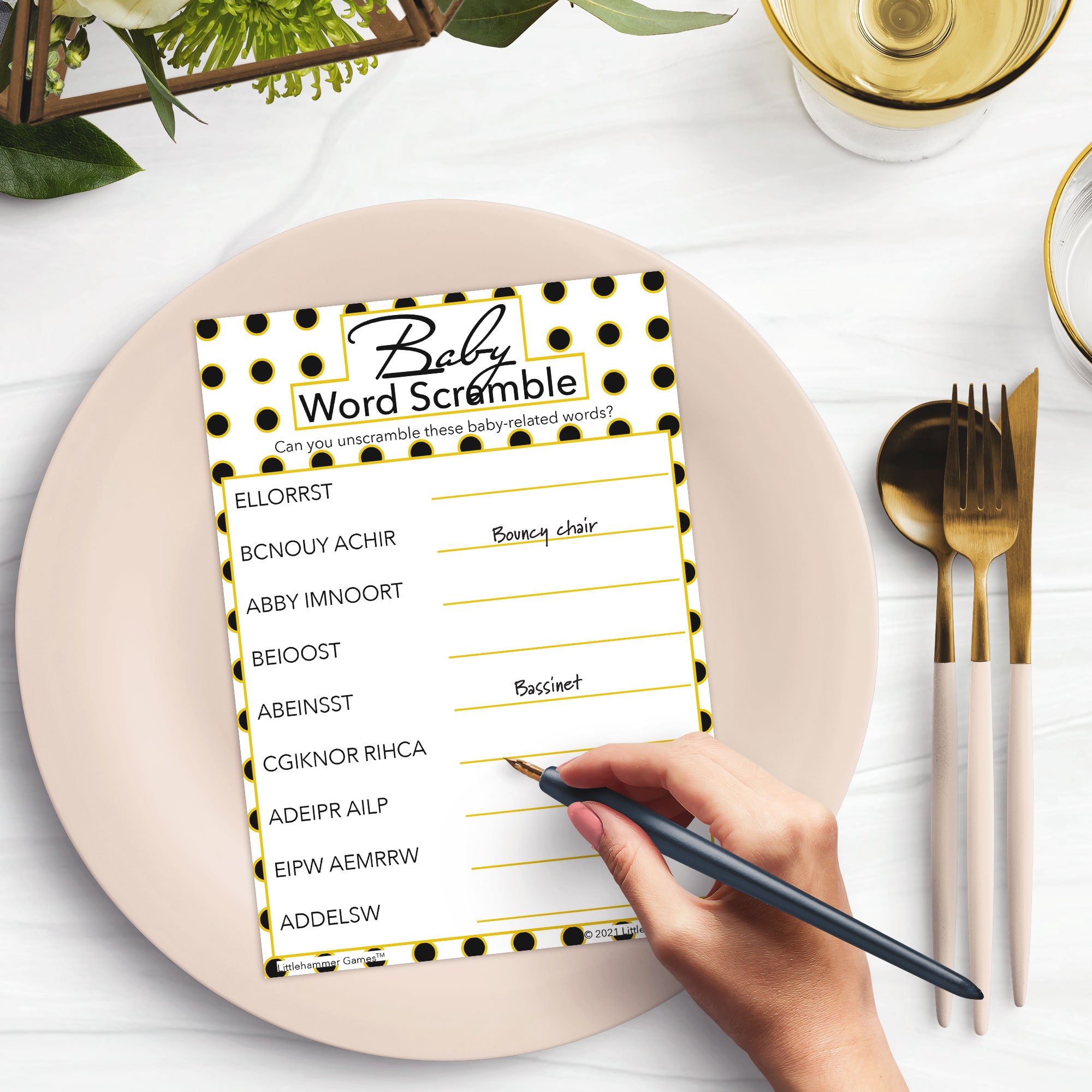 Woman’s hand holding a pen playing a geometric black and gold polka dot Baby Word Scramble game card at a light place setting
