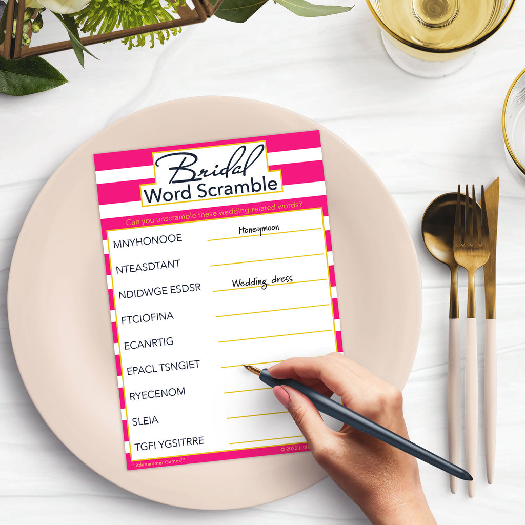 Woman’s hand holding a pen playing a pink-striped Bridal Word Scramble game card at a light place setting