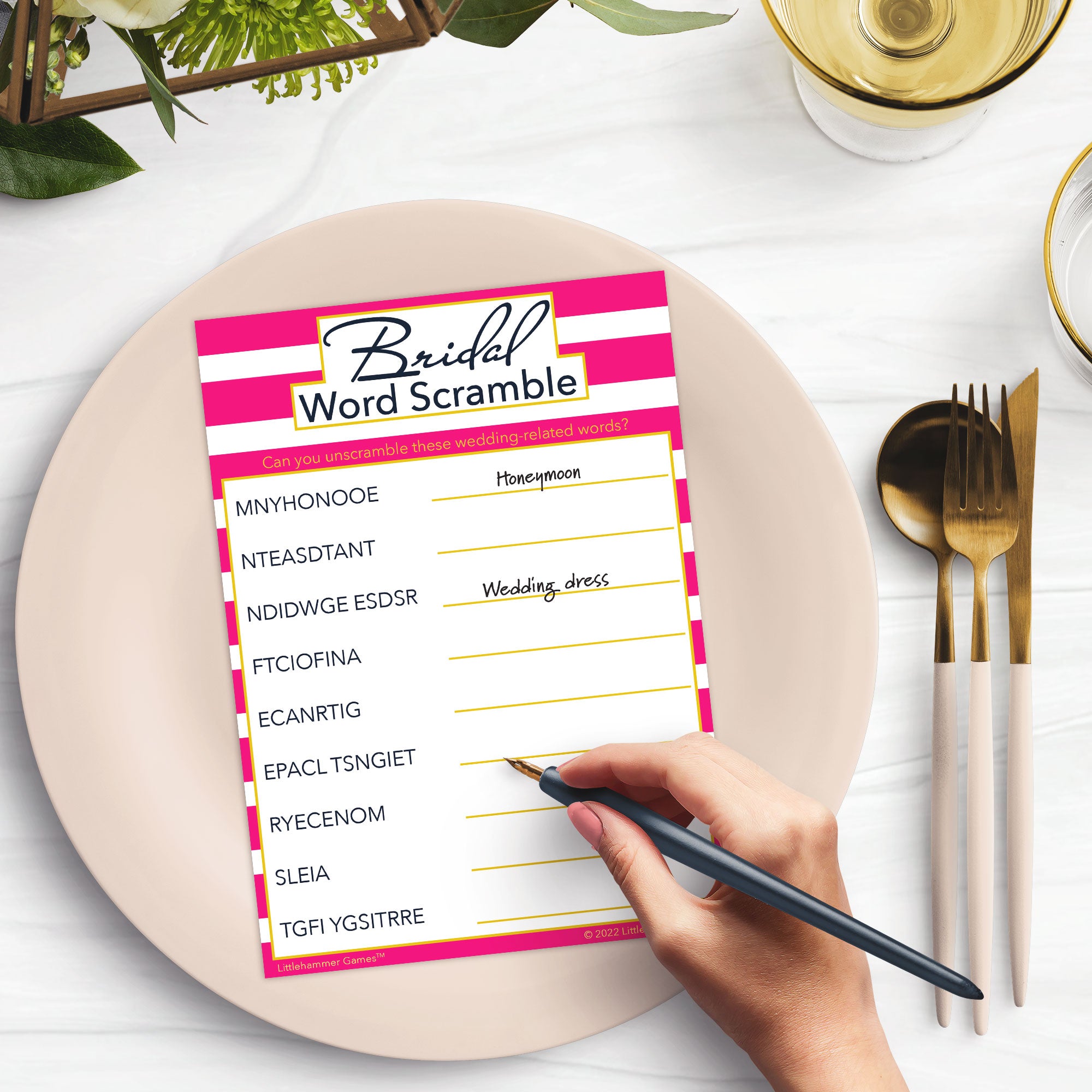 Woman’s hand holding a pen playing a pink-striped Bridal Word Scramble game card at a light place setting