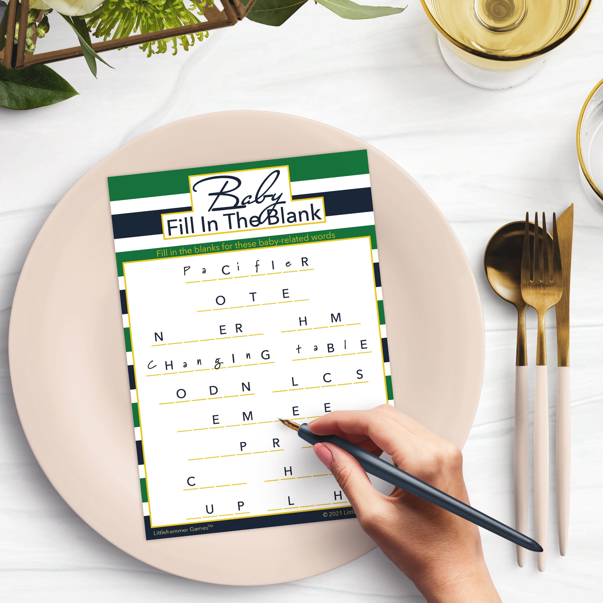 Woman’s hand holding a pen playing a green and navy-striped Baby Fill in the Blank game card at a light place setting