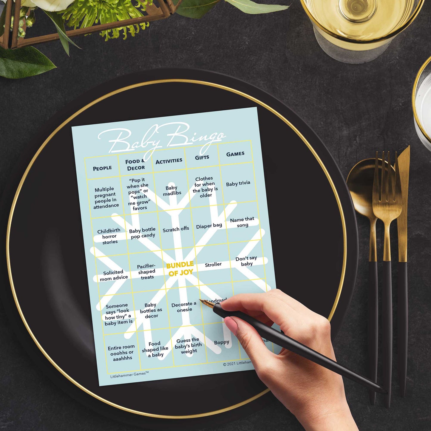 Woman with a pen sitting at a table with a snowflake-themed Baby Bingo game card on a black and gold plate at a dark place setting