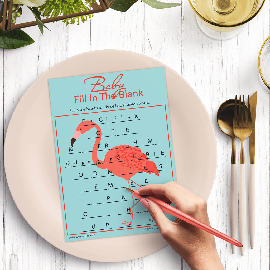Woman’s hand holding a pen playing a flamingo-themed Baby Fill in the Blank game card at a light place setting