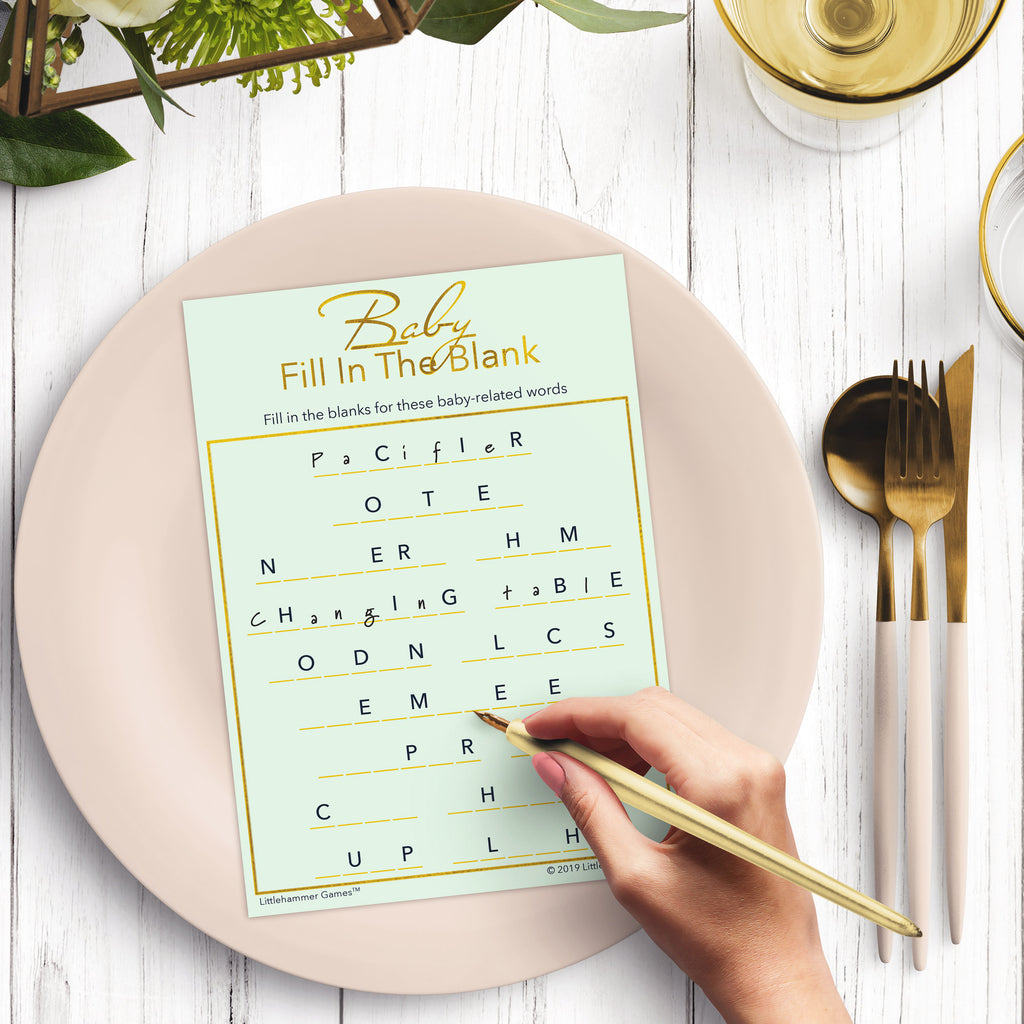 Woman’s hand holding a pen playing a ming and gold Baby Fill in the Blank game card at a light place setting