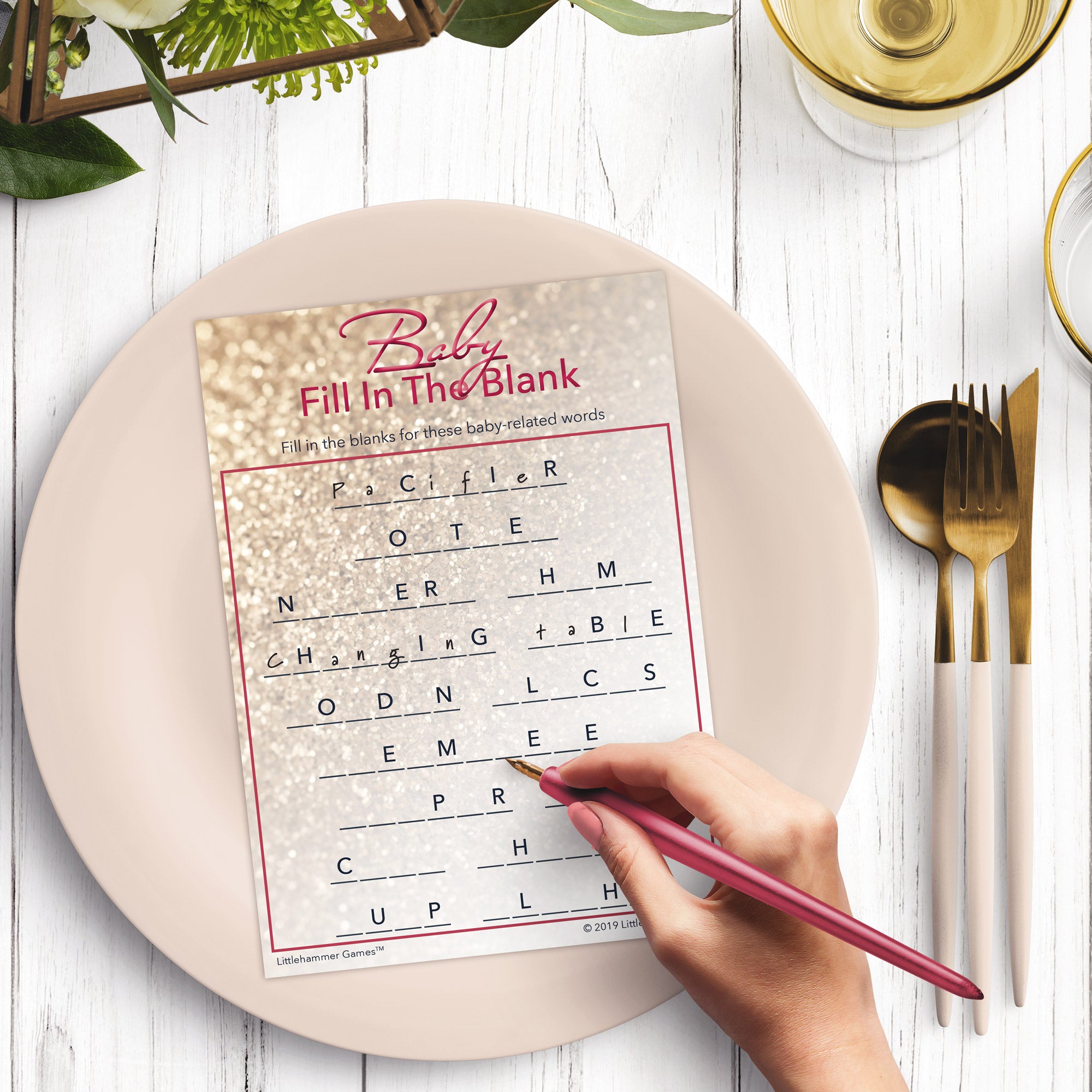 Woman’s hand holding a pen playing a glittery rose gold Baby Fill in the Blank game card at a light place setting
