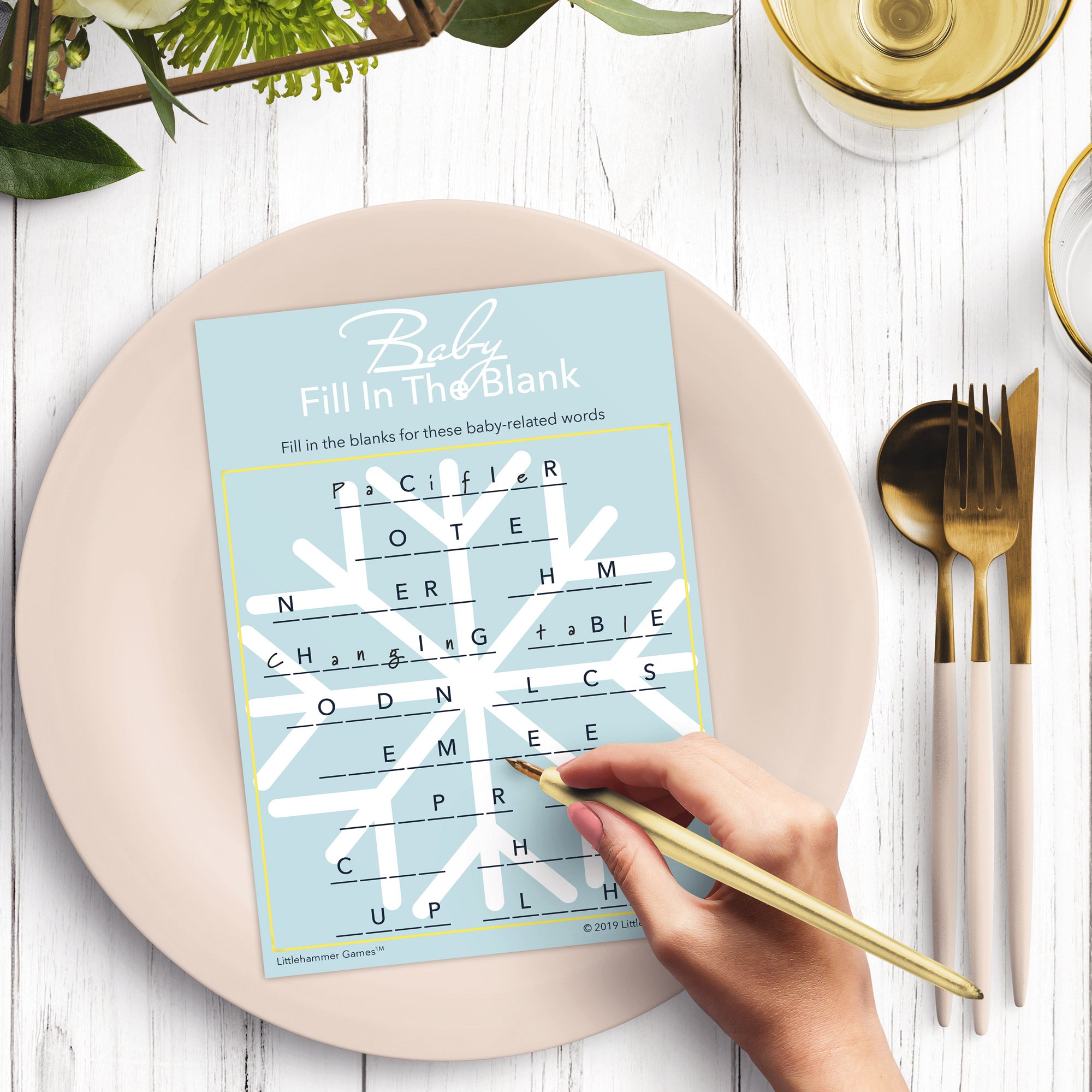Woman’s hand holding a pen playing a snowflake design Baby Fill in the Blank game card at a light place setting