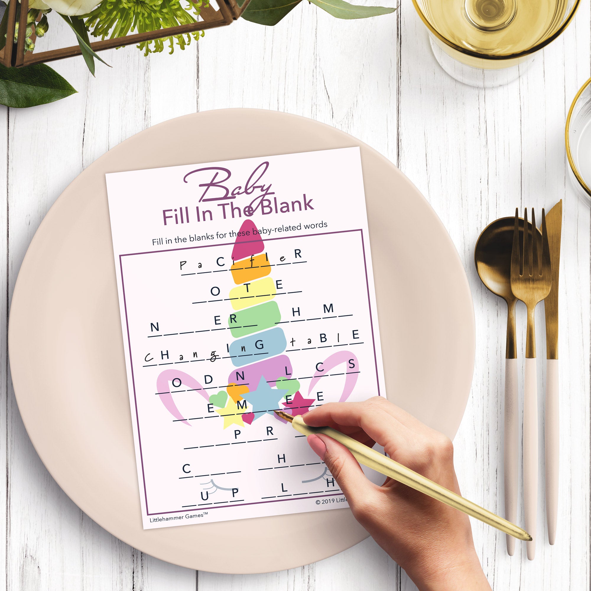 Woman’s hand holding a pen playing a unicorn theme Baby Fill in the Blank game card at a light place setting