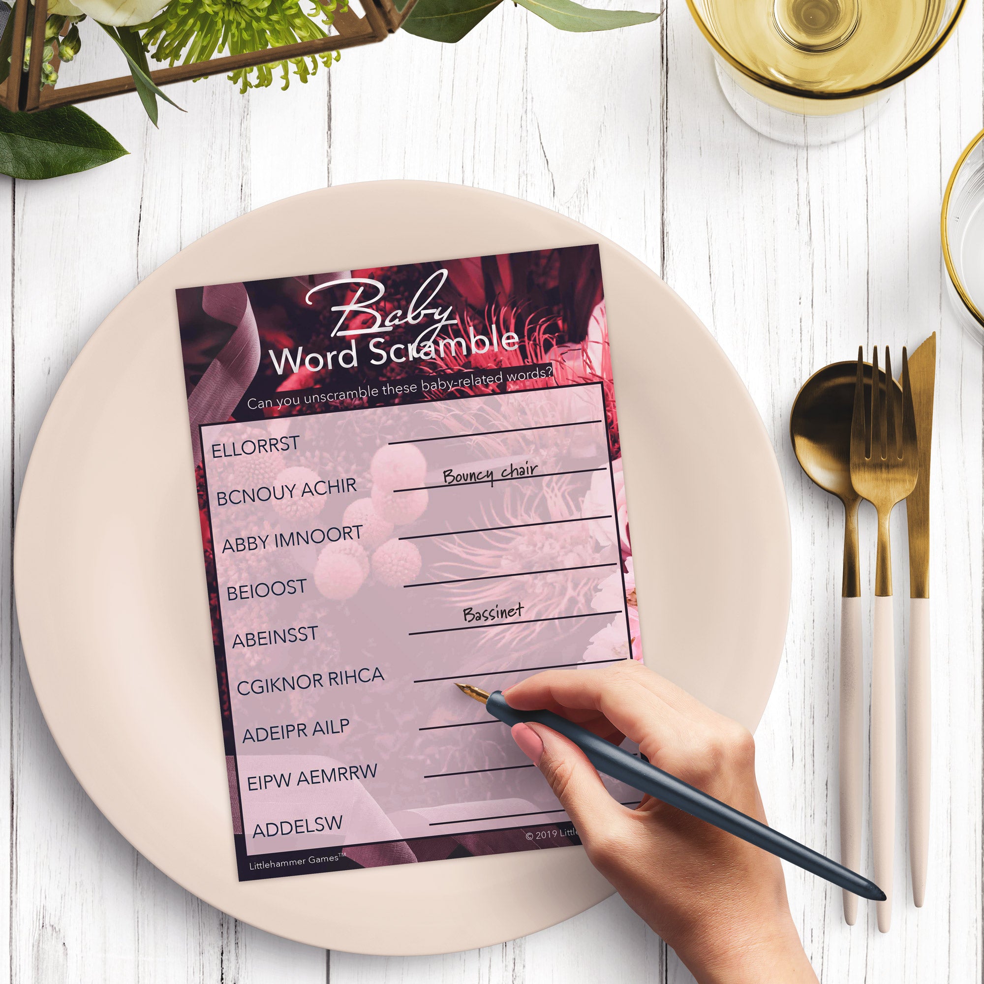 Woman’s hand holding a pen playing a purple floral Baby Word Scramble game card at a light place setting