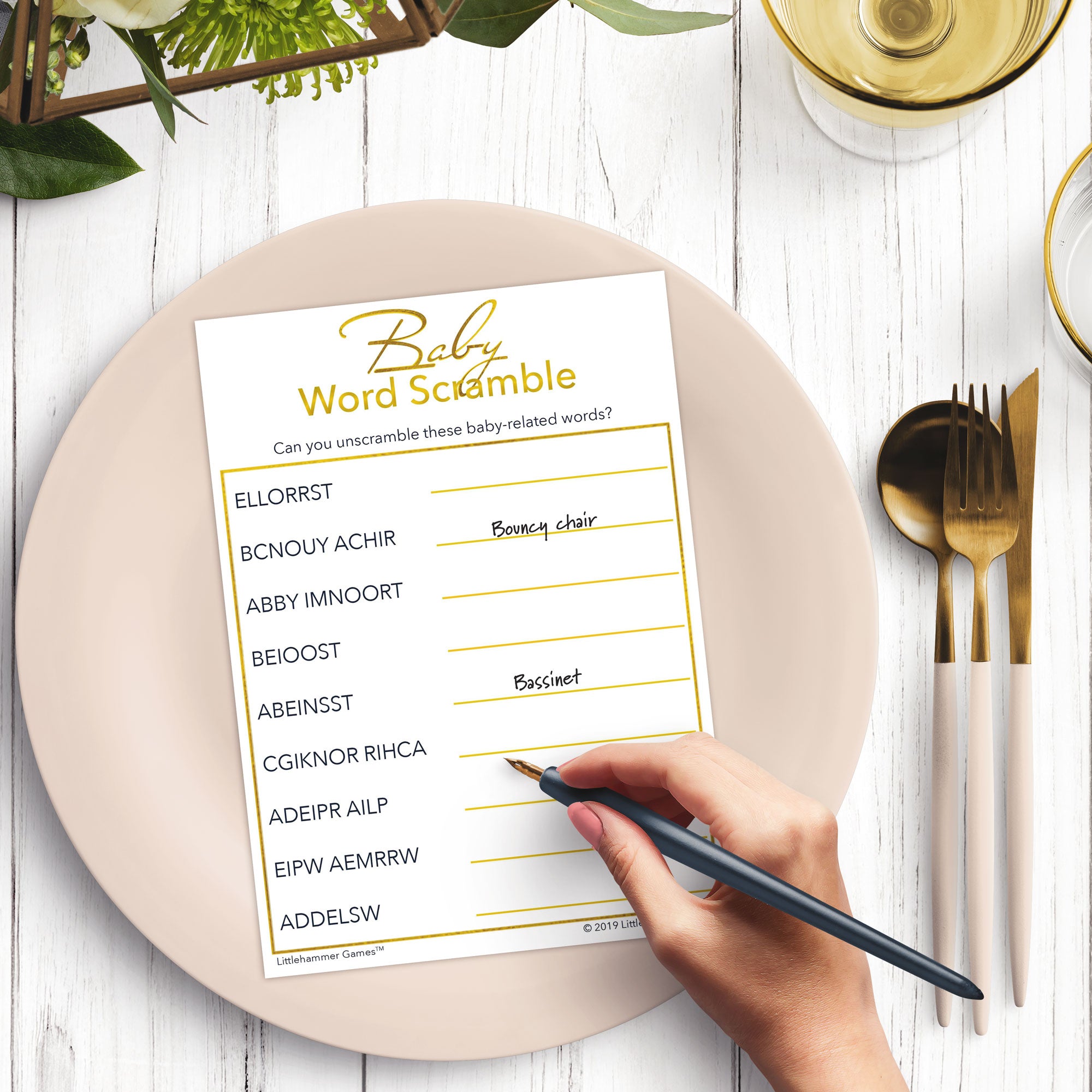 Woman’s hand holding a pen playing a minimalist gold and white Baby Word Scramble game card at a light place setting