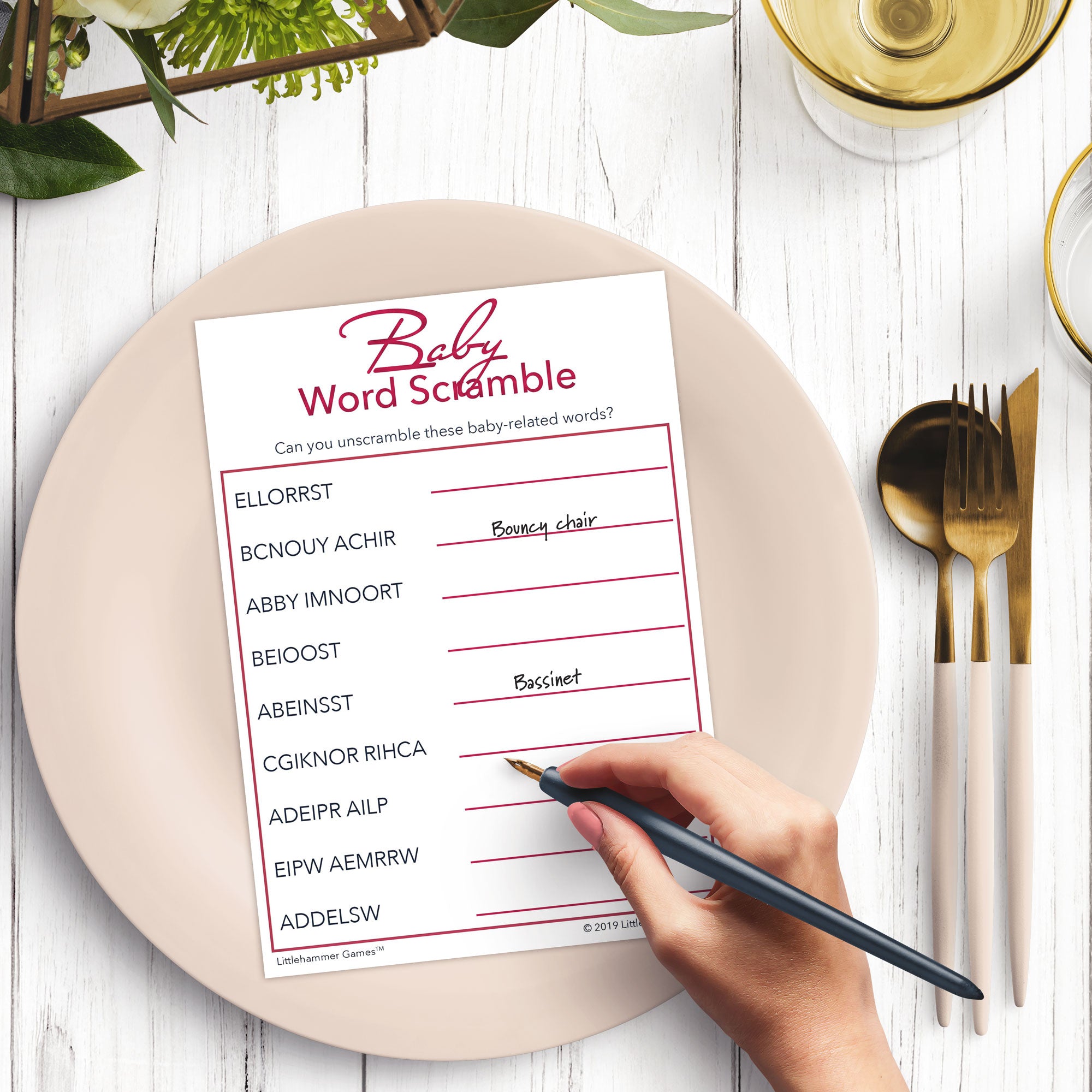 Woman’s hand holding a pen playing a rose gold and white Baby Word Scramble game card at a light place setting
