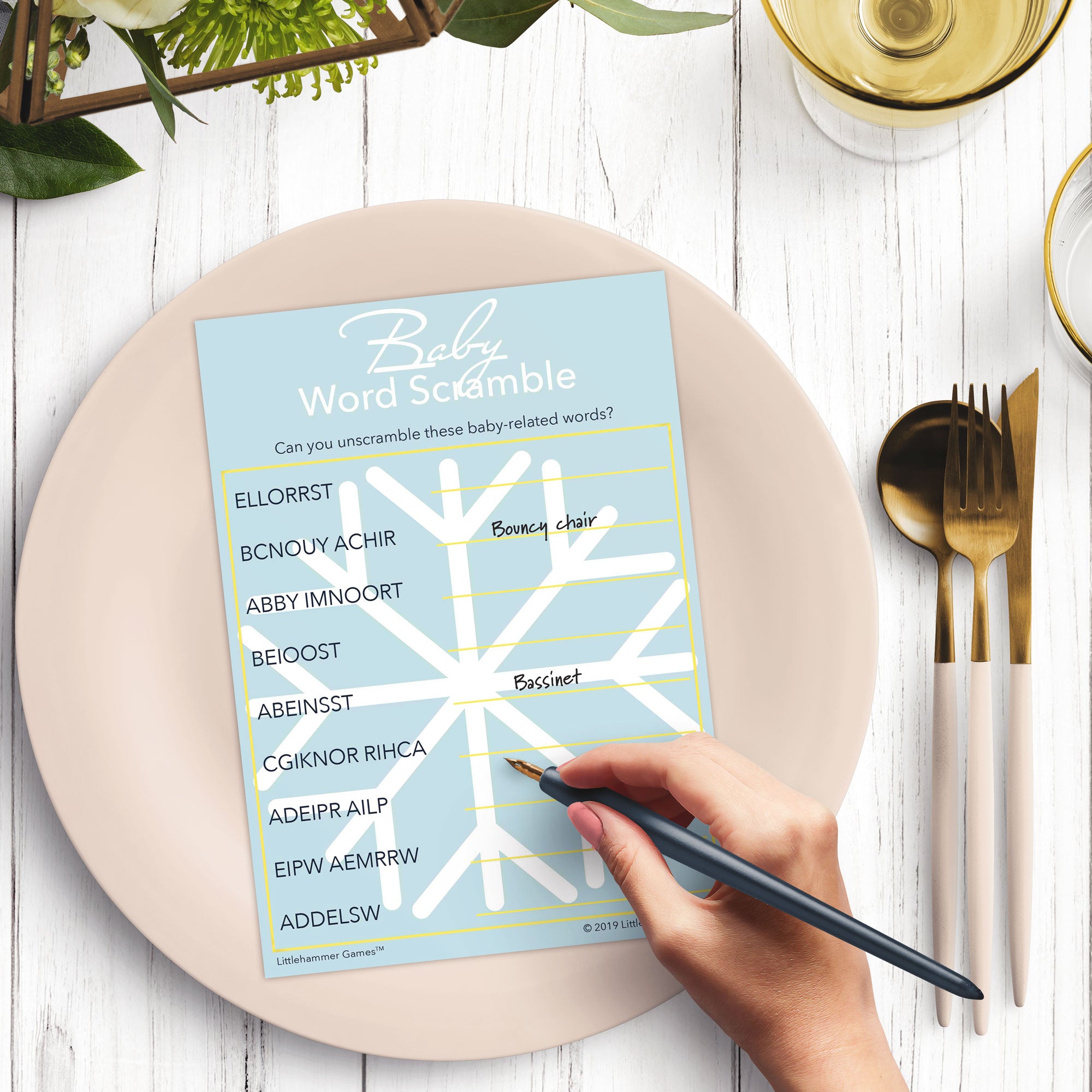 Woman’s hand holding a pen playing a pale snowflake-themed Baby Word Scramble game card at a light place setting