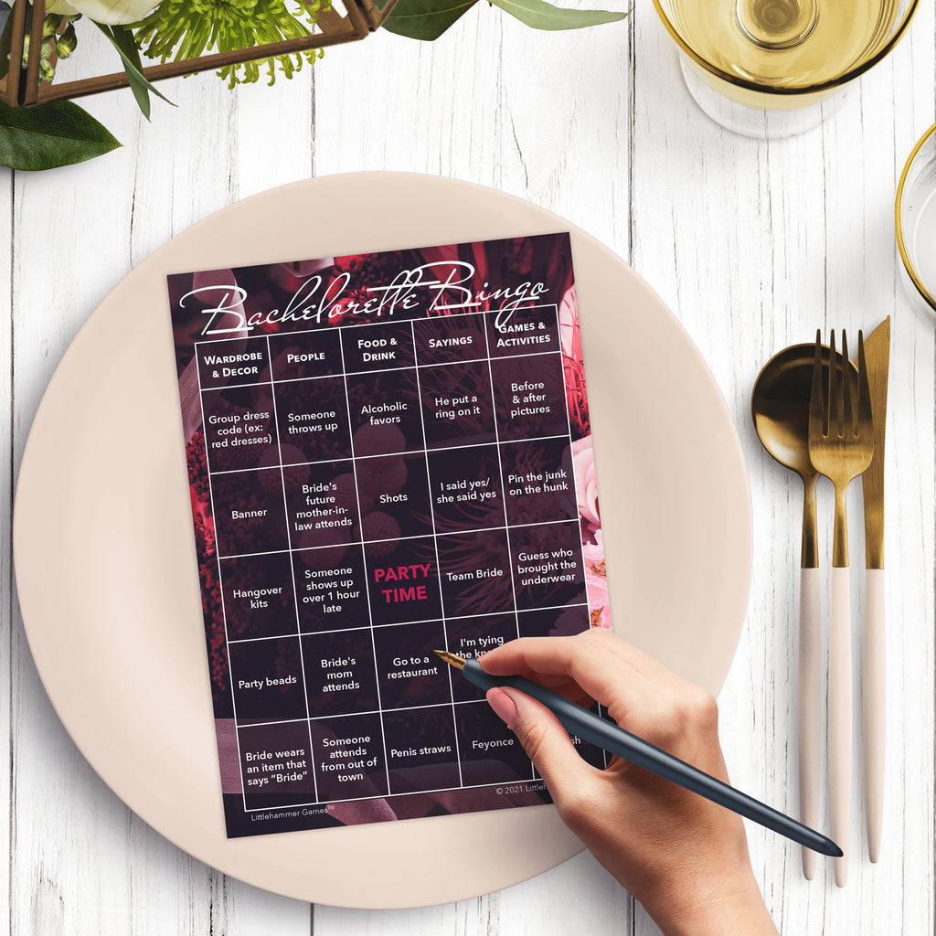 Woman’s hand holding a pen playing a floral design Bachelorette Bingo game card at a light place setting