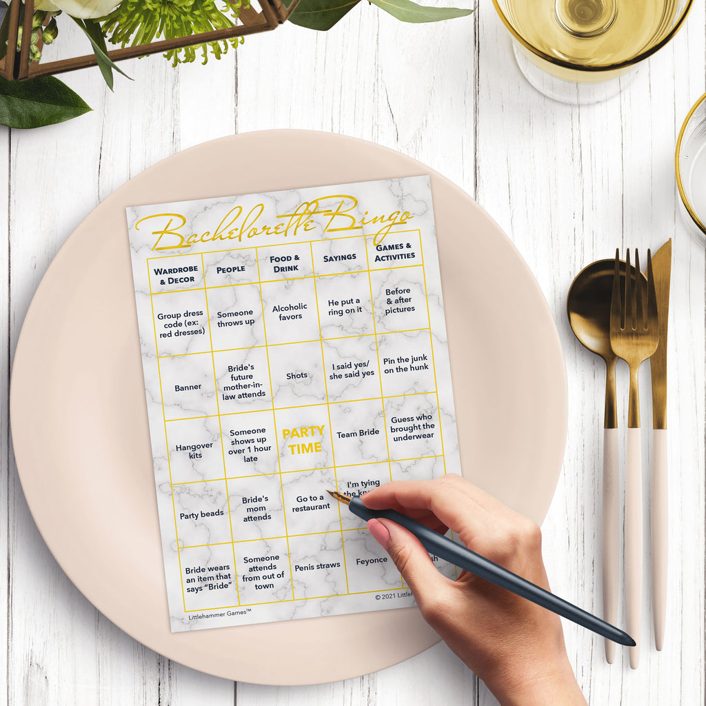 Woman’s hand holding a pen playing a gold and marble Bachelorette Bingo game card at a light place setting