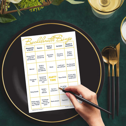 Woman with a pen sitting at a table with a gold and white Bachelorette Bingo game card on a gold and black plate on a dark place setting