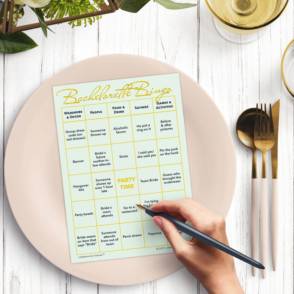 Woman’s hand holding a pen playing a mint and gold Bachelorette Bingo game card at a light place setting