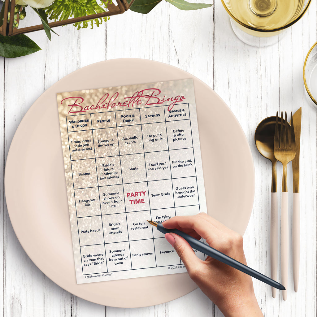 Woman’s hand holding a pen playing a glittery rose gold Bachelorette Bingo game card at a light place setting