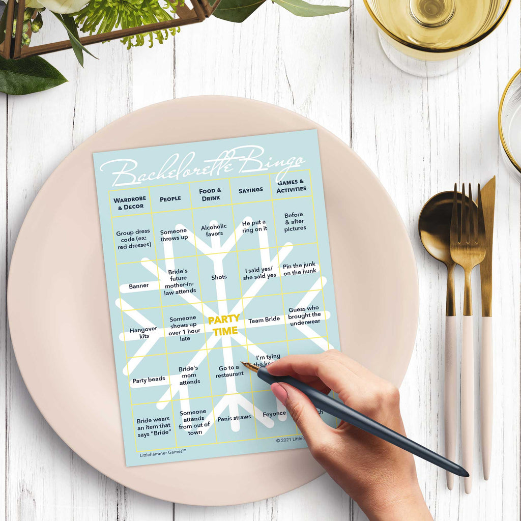 Woman’s hand holding a pen playing a snowlfake-themed Bachelorette Bingo game card at a light place setting