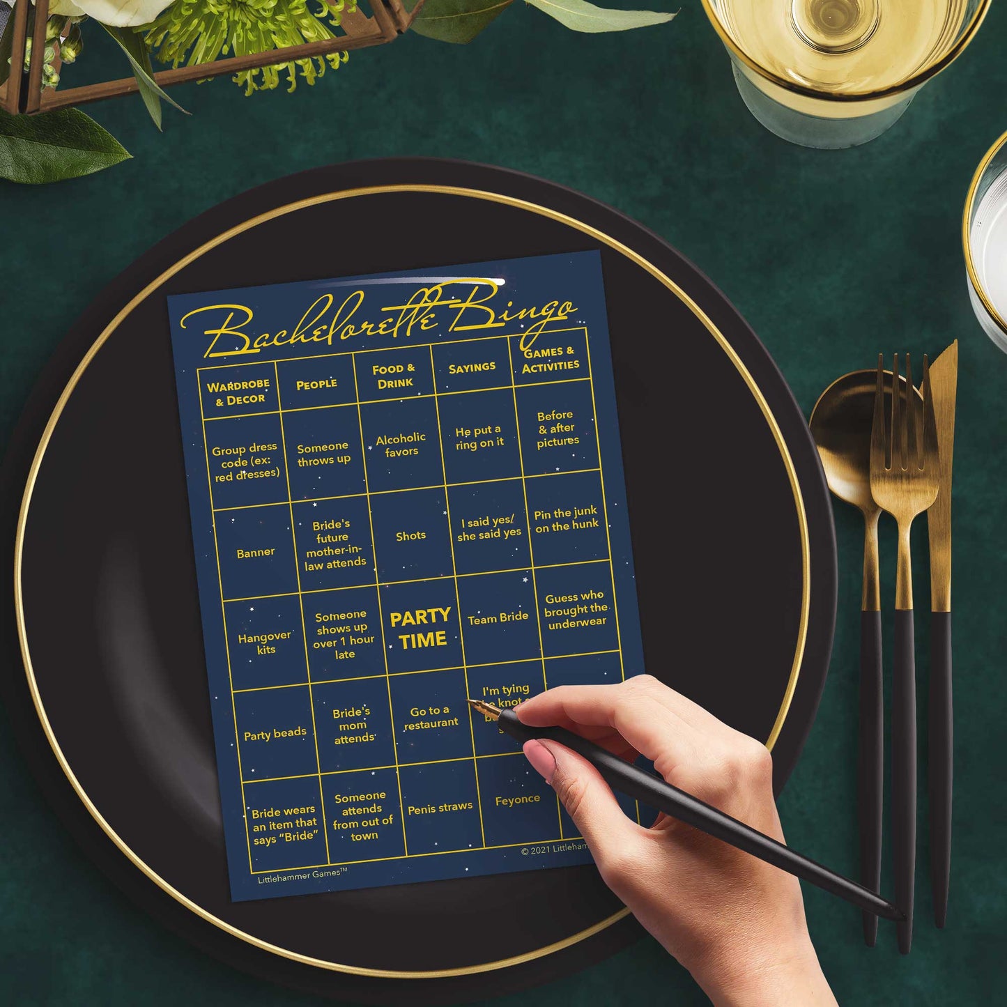 Woman with a pen sitting at a table with a celestial themed Bachelorette Bingo game card on a gold and black plate on a dark place setting