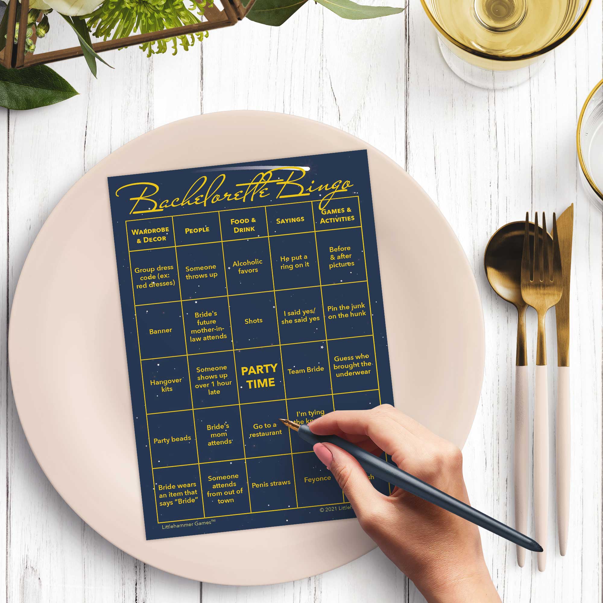 Woman’s hand holding a pen playing a celestial Bachelorette Bingo game card at a light place setting