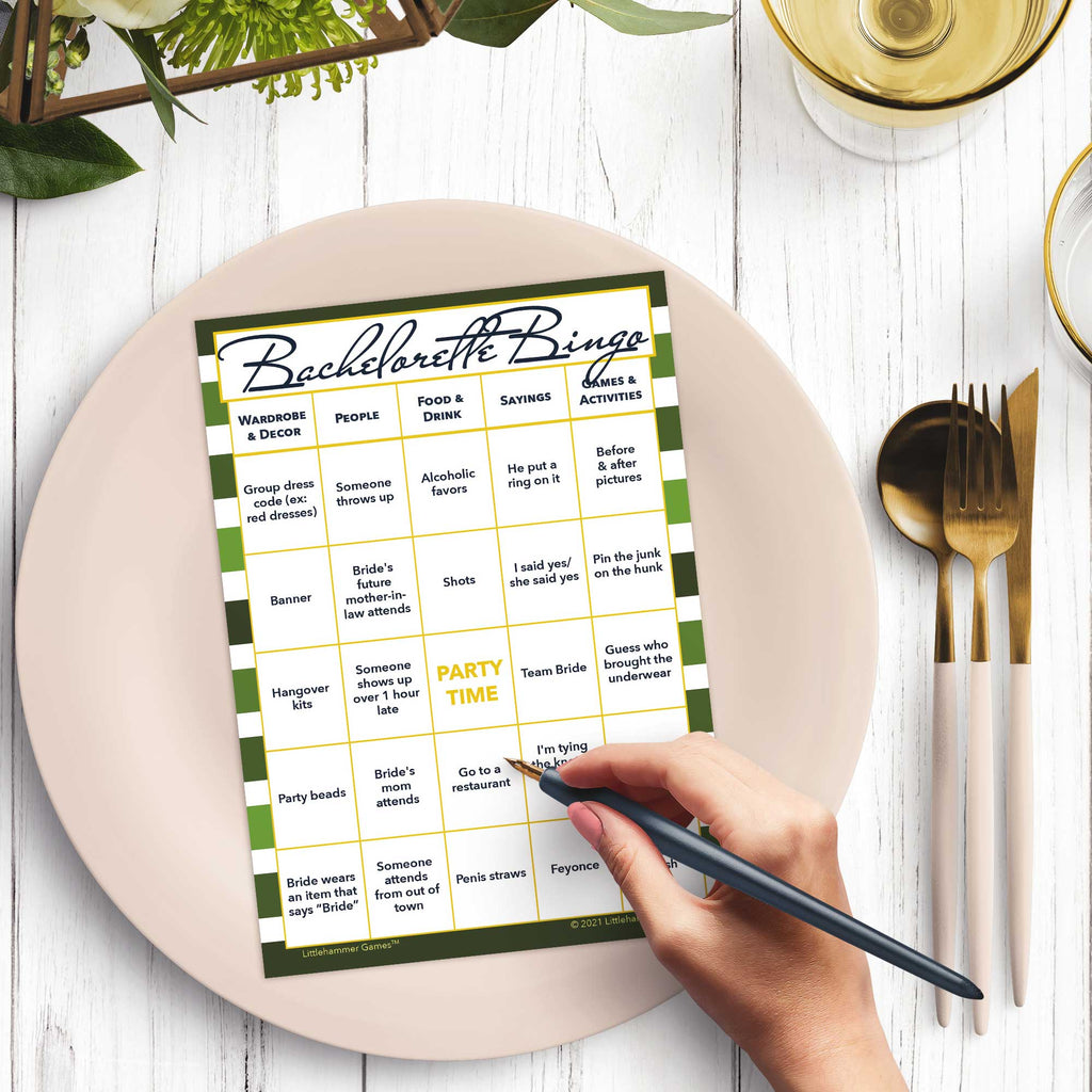 Woman’s hand holding a pen playing a shades of green stripes Bachelorette Bingo game card at a light place setting