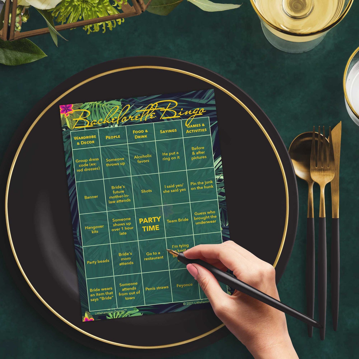 Woman with a pen sitting at a table with a tropical themed Bachelorette Bingo game card on a gold and black plate on a dark place setting