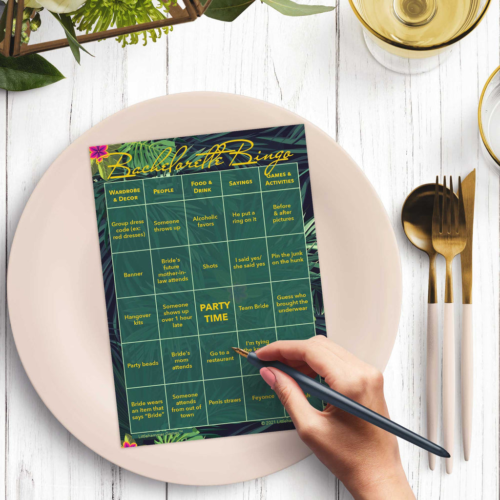 Woman’s hand holding a pen playing a jungle leaves Bachelorette Bingo game card at a light place setting