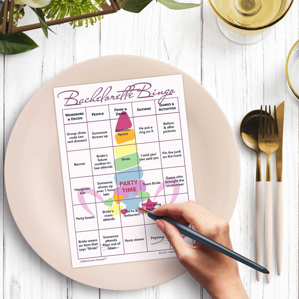 Woman’s hand holding a pen playing a unicorn-themed Bachelorette Bingo game card at a light place setting