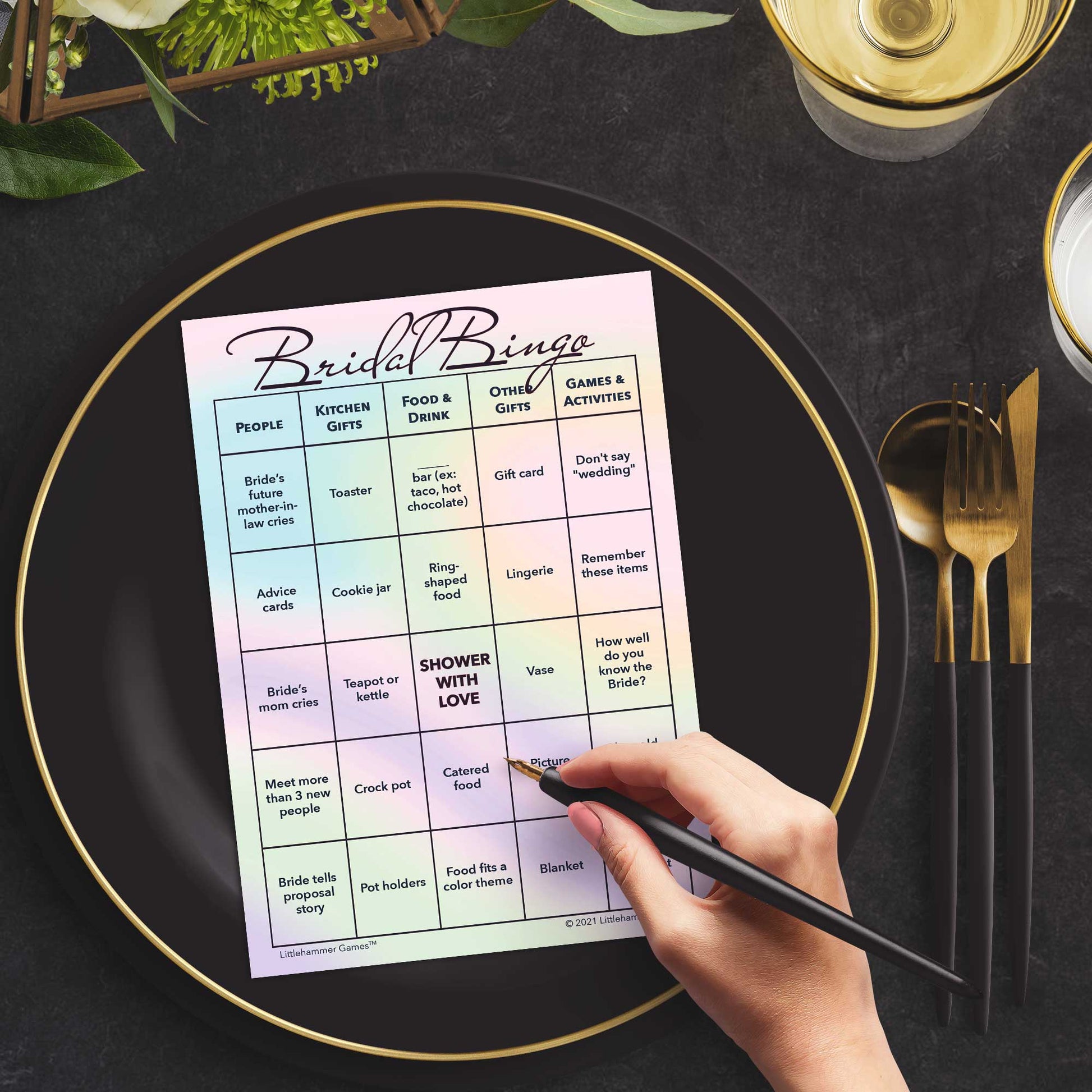 Woman with a pen sitting at a table with a hologram-themed Bridal Bingo game card on a black and gold plate at a dark place setting