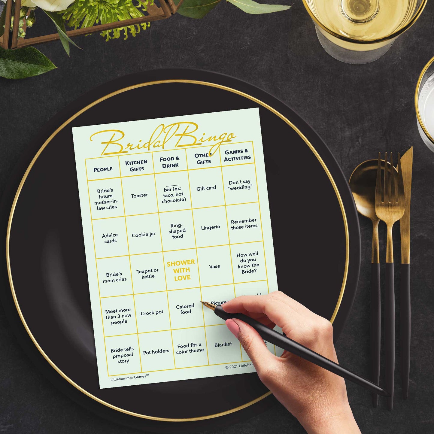 Woman with a pen sitting at a table with a mint and gold Bridal Bingo game card on a black and gold plate at a dark place setting