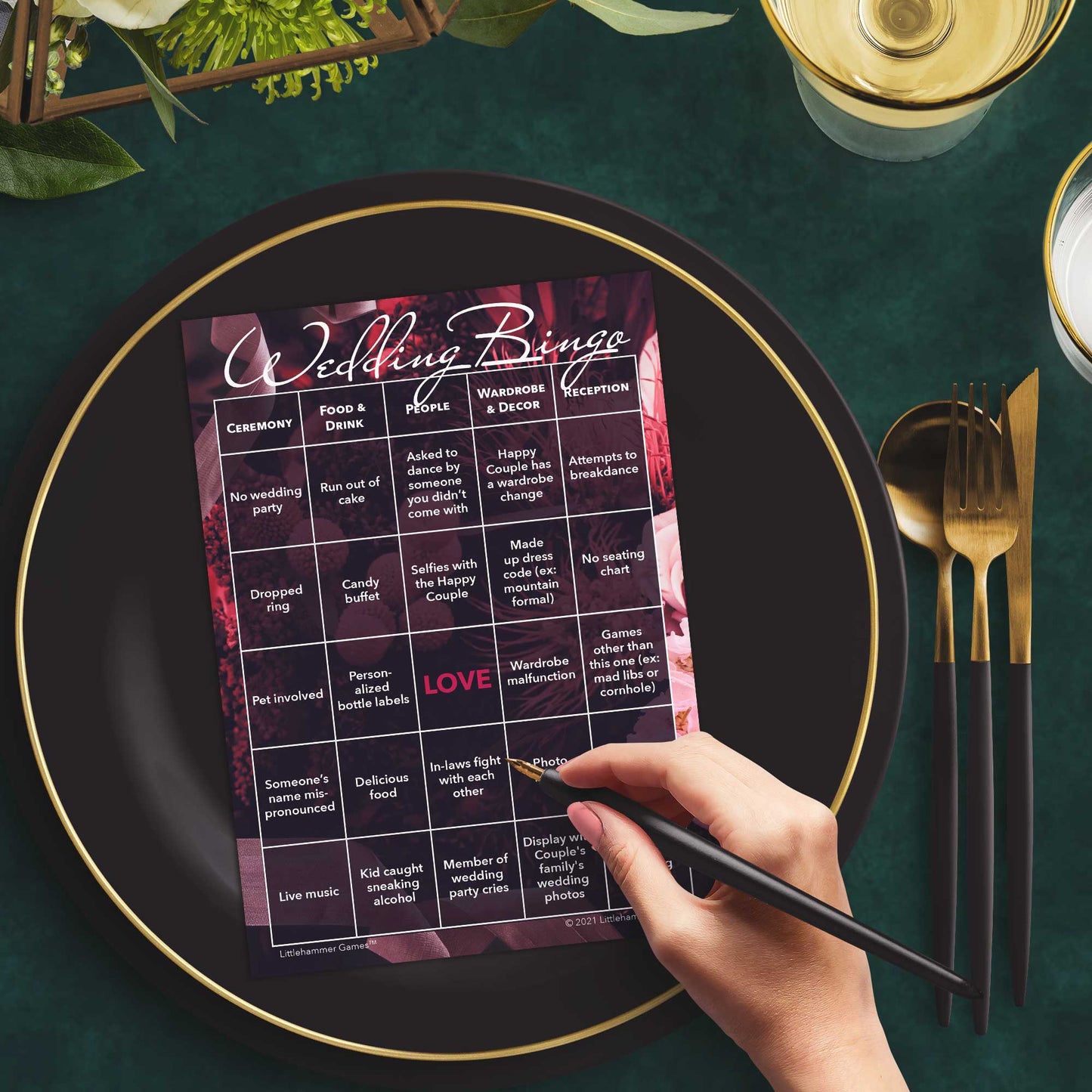 Woman with a pen sitting at a table with a dark floral Wedding Bingo game card on a black and gold plate at a dark place setting