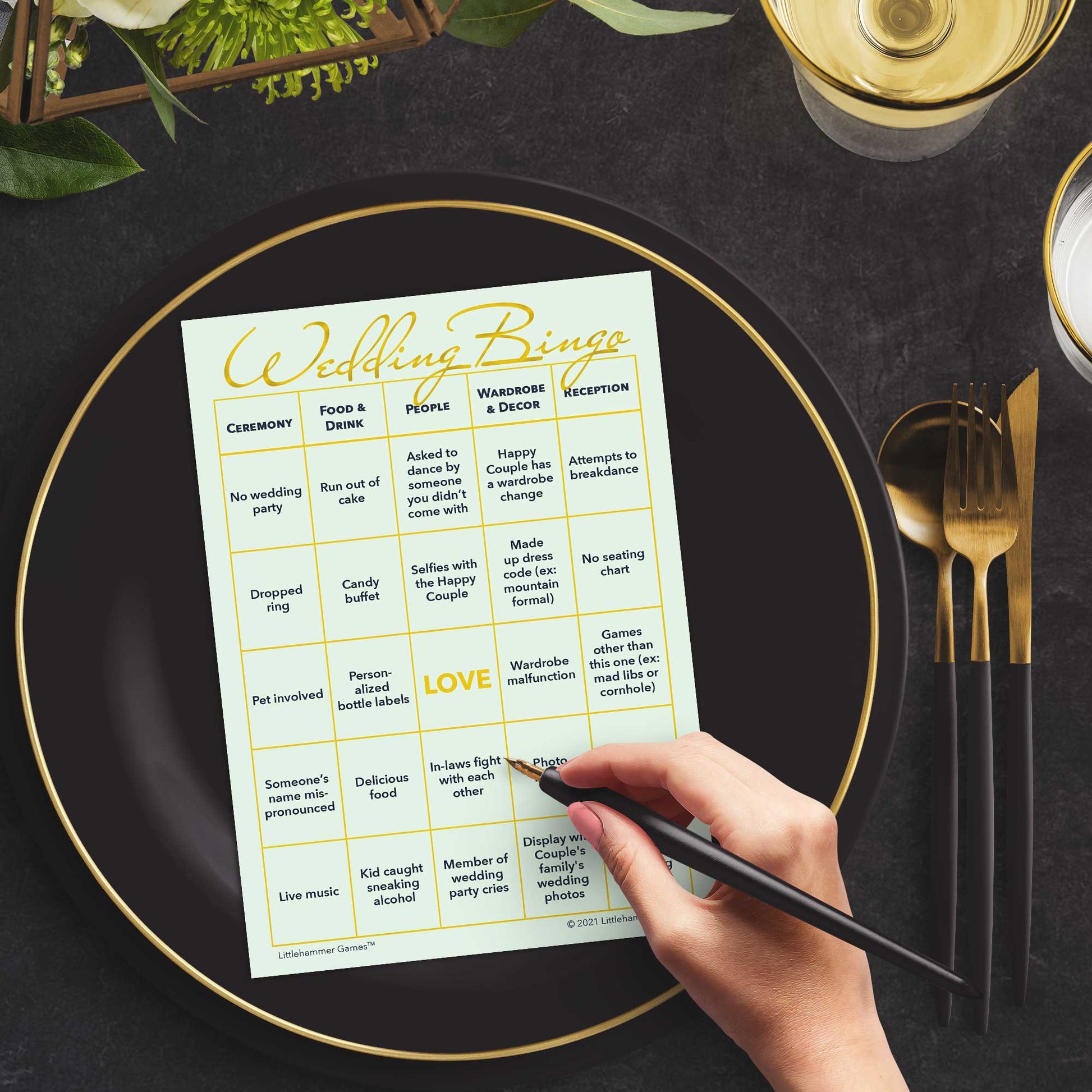 Woman with a pen sitting at a table with a mint and gold Wedding Bingo game card on a black and gold plate at a dark place setting